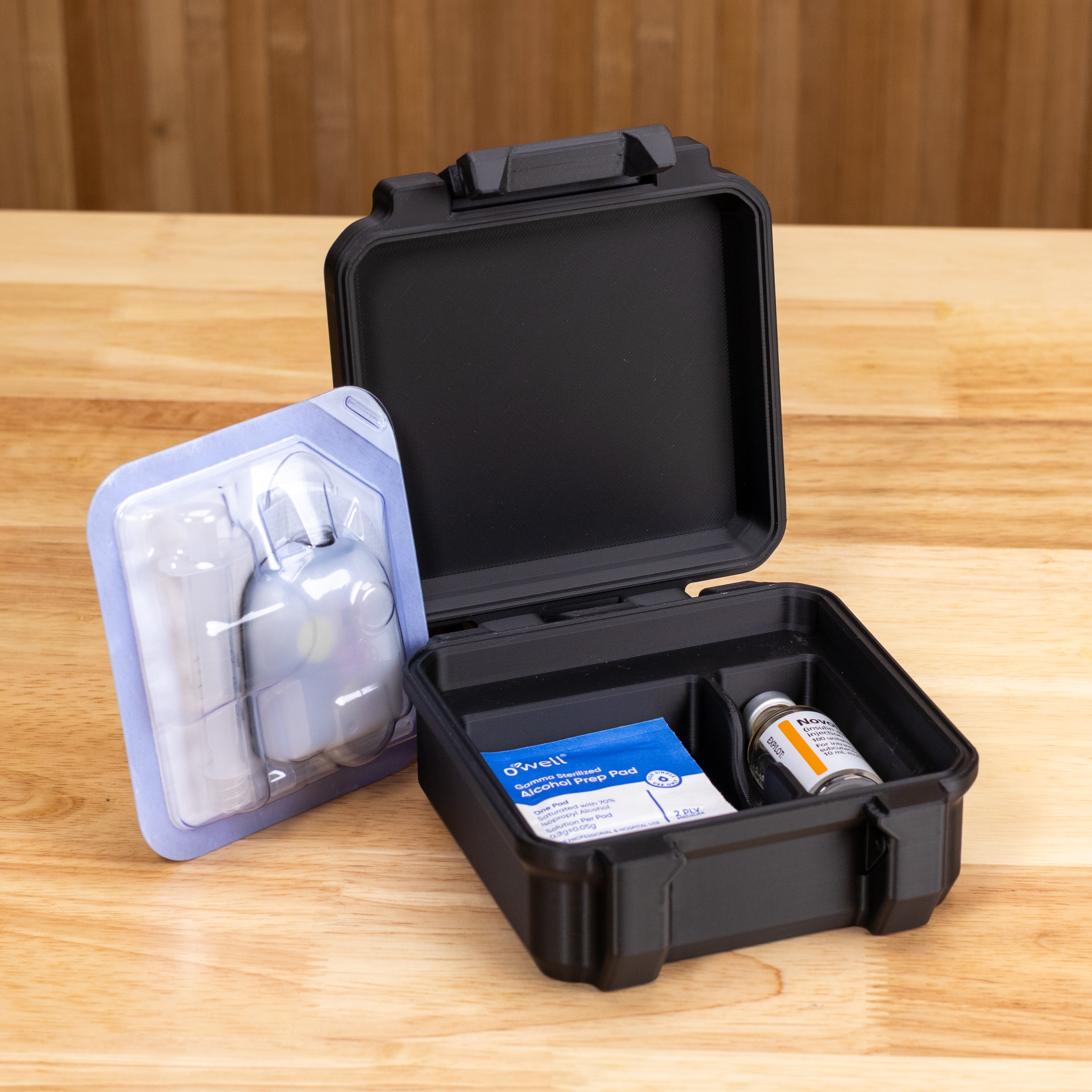 An open black hard-sided medical storage case on a light wood tabletop, with a blister-pack accessory leaning against it. Visible contents include a small blue-and-white box and a vial.